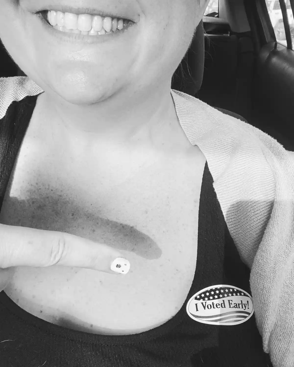 A black-and-white photo shows a smiling person in a car, wearing a black top with a white cardigan, pointing to a small…