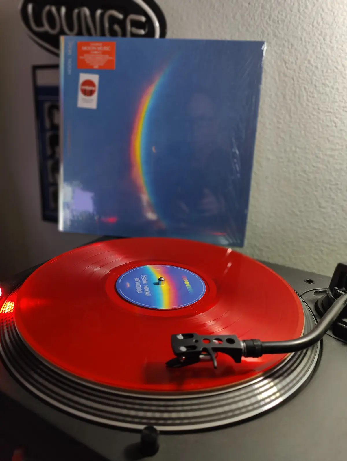 A red vinyl record spins on a turntable with its album cover standing upright behind it, featuring a blue cover with a…