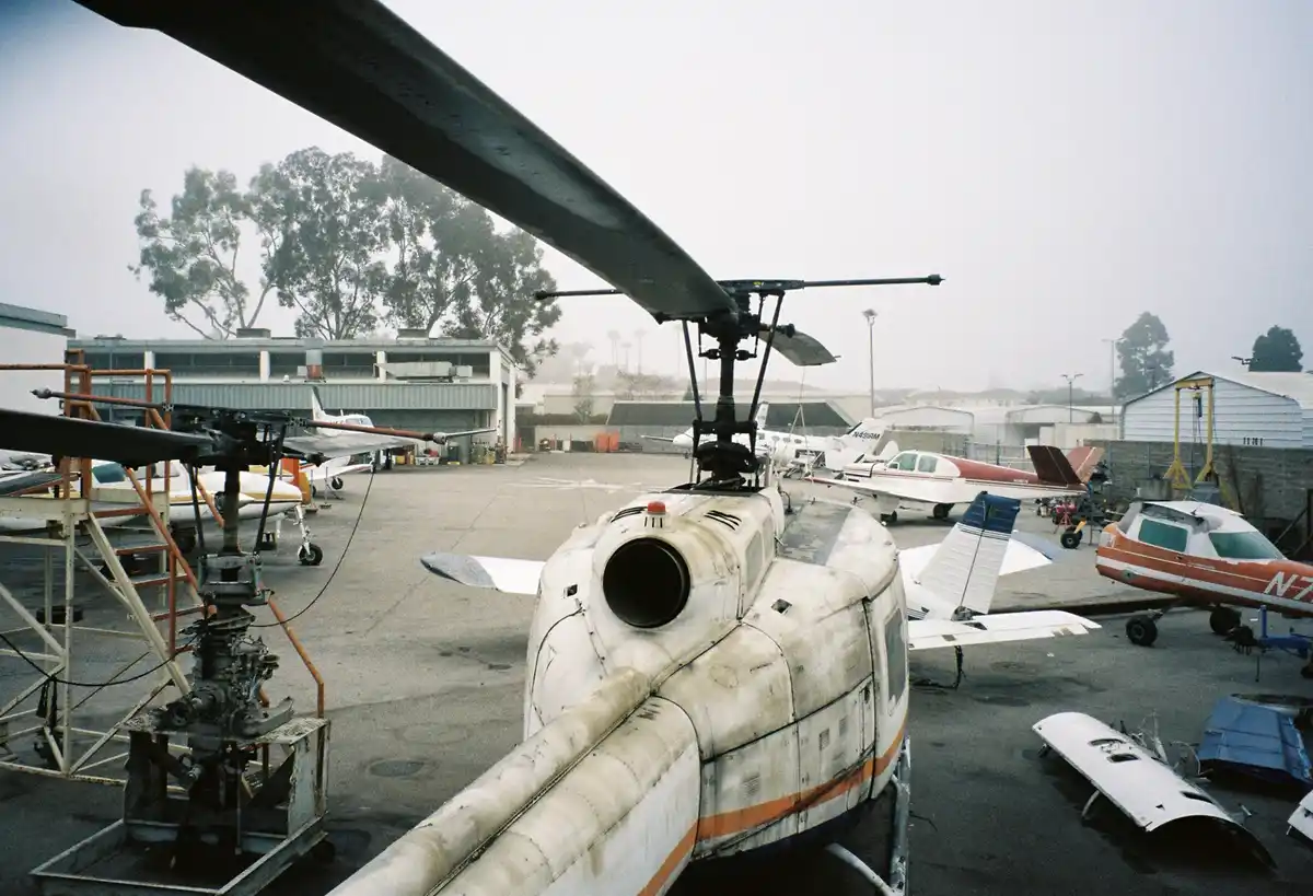 A weathered helicopter sits in an aircraft hangar yard, surrounded by other small planes and maintenance equipment. The…