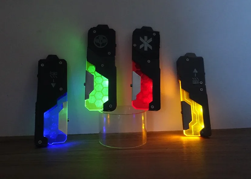 Four black, angular, futuristic-looking devices with glowing panels in blue, green, red, and yellow stand on a wooden…
