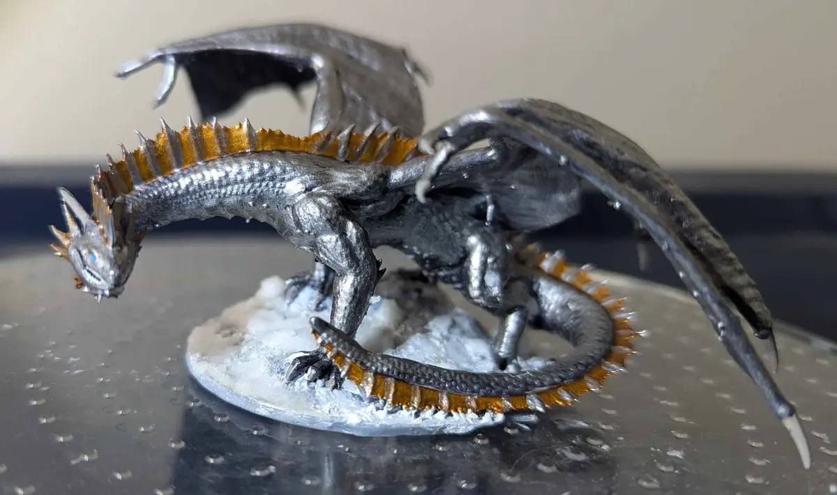 A silver-painted dragon figurine with gold-tipped spikes and wings is posed on a snowy base, looking down with a stern…