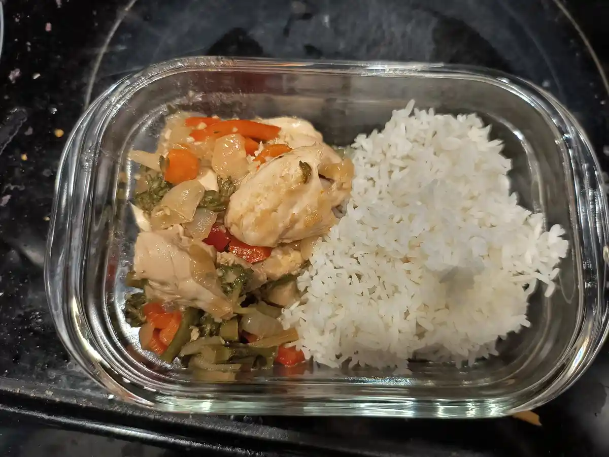 A clear glass meal prep container holds a portion of white rice next to a serving of cooked chicken mixed with chopped…