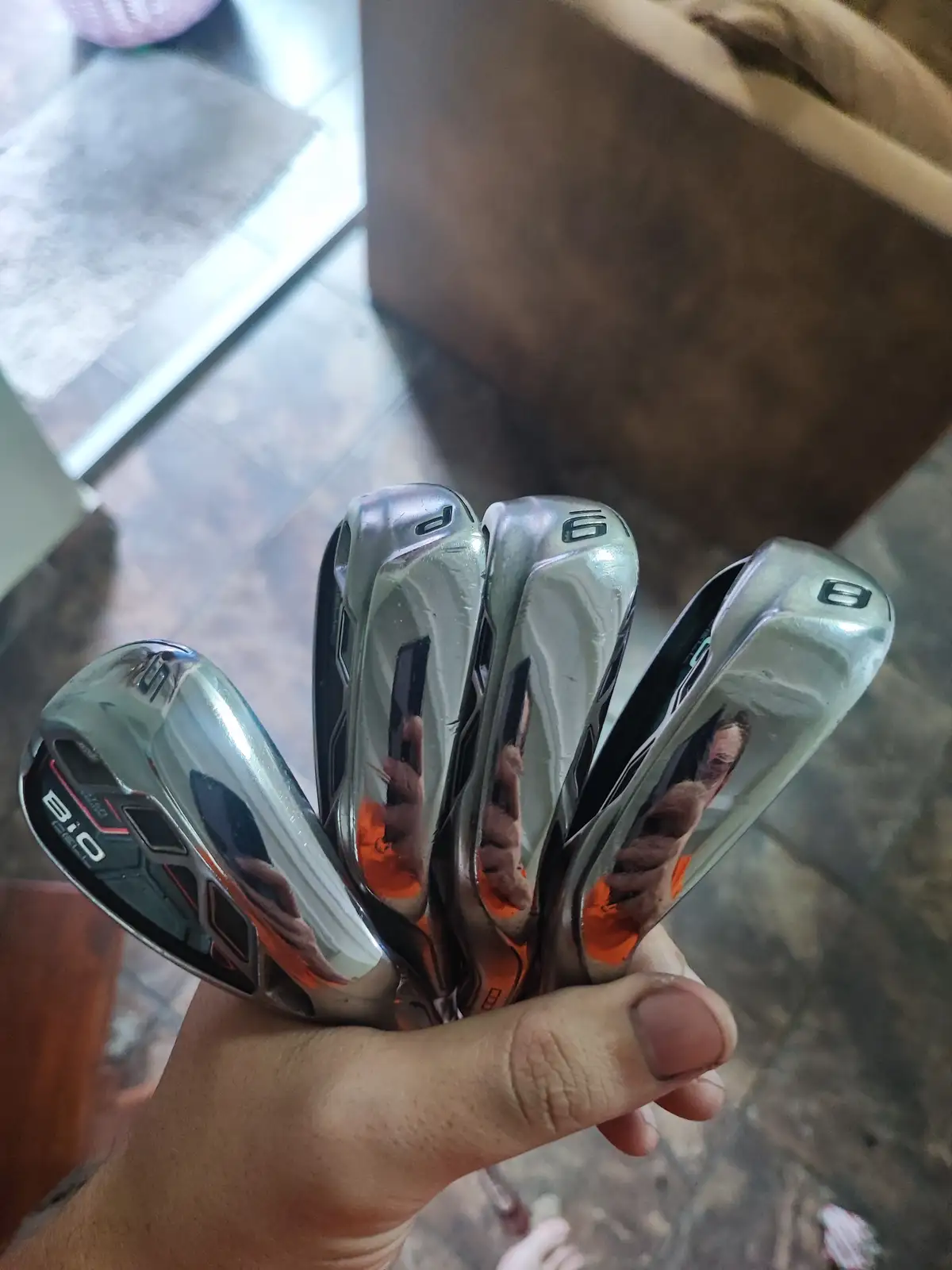 A hand holds a fan of shiny, chrome-finished golf irons, showing club numbers 6, 7, 8, and 9, with the 'BIO' brand visible…