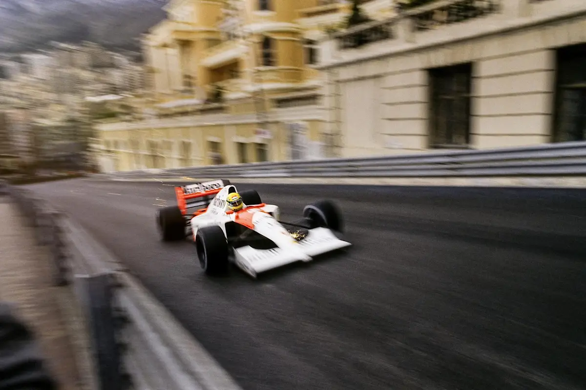 A dynamic, motion-blurred photograph captures a white and orange Formula One race car speeding along a narrow city street…