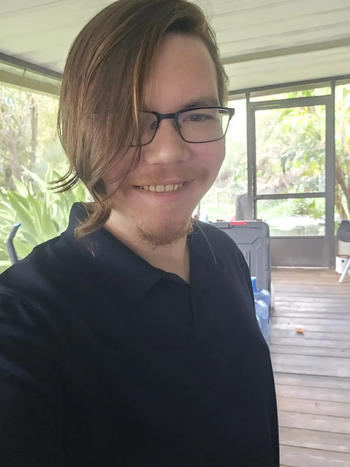 A person with shoulder-length brown hair and glasses smiles at the camera while wearing a black polo shirt, standing on a…