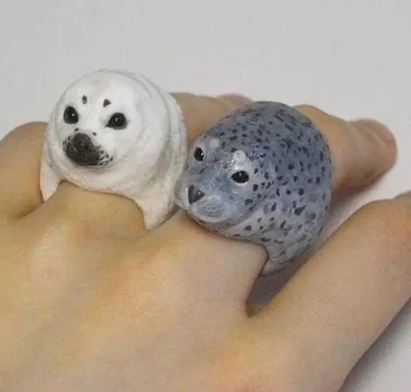 Two small, realistic-looking seal figurines are worn as rings on a person's fingers, one white and one spotted gray, both…