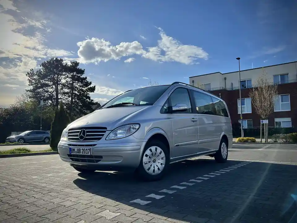 A silver Mercedes-Benz Viano minivan is parked on a paved lot under a blue sky with scattered clouds. The car's German…
