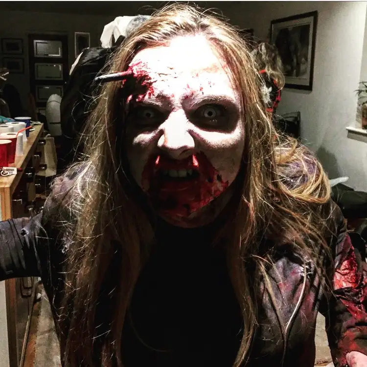A person with long hair, wearing a dark jacket, is shown with heavy zombie makeup including fake blood, white face paint…