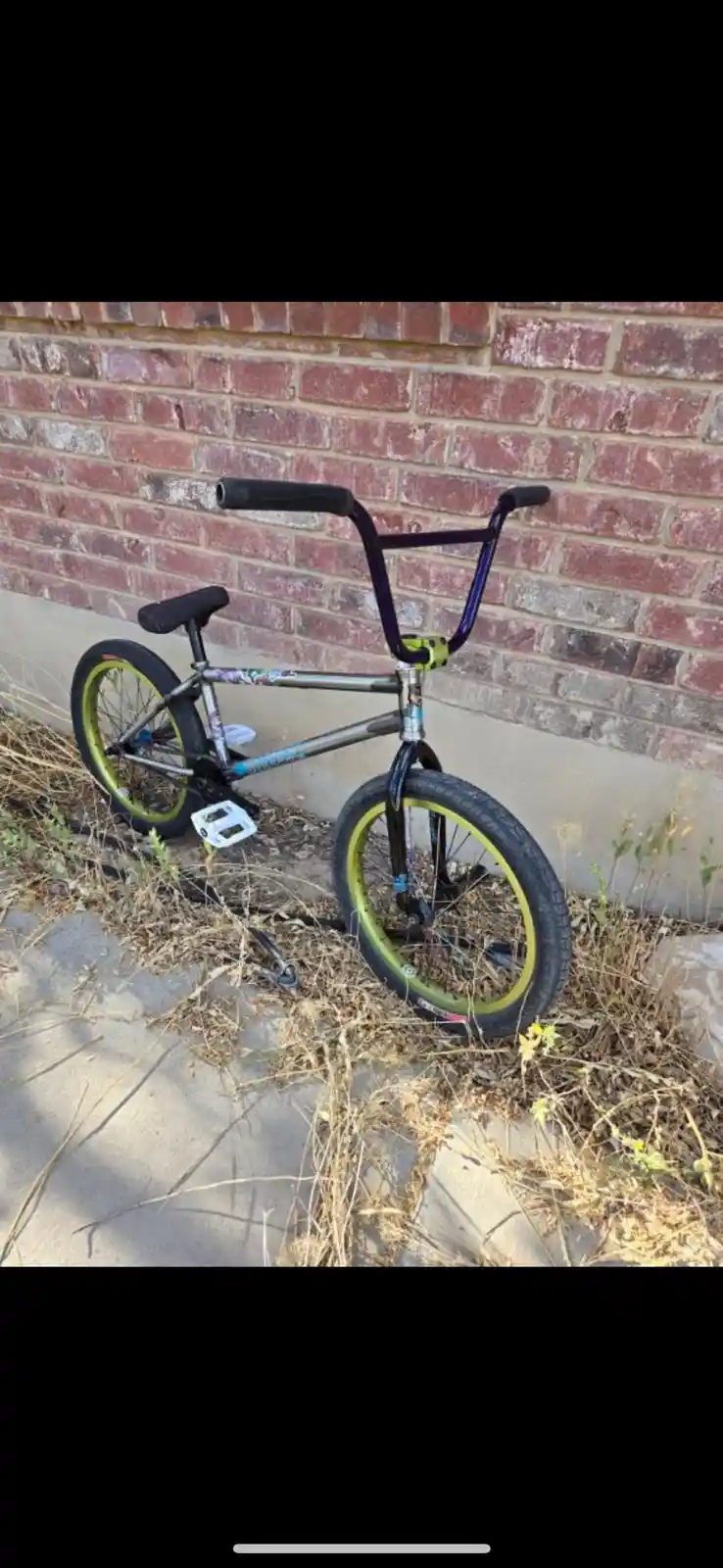 A BMX bike with purple handlebars and yellow rims leans against a red brick wall next to overgrown dry grass and a concrete…
