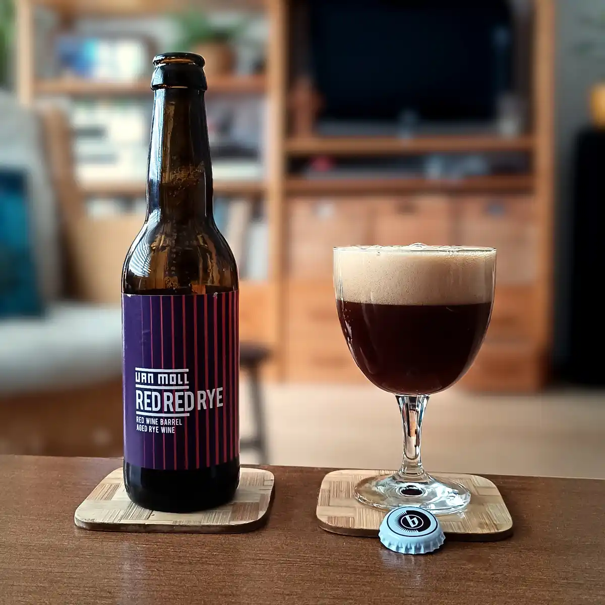 A brown bottle of Van Moll Red Red Rye beer sits next to a glass filled with dark beer and a thick white head, both resting…