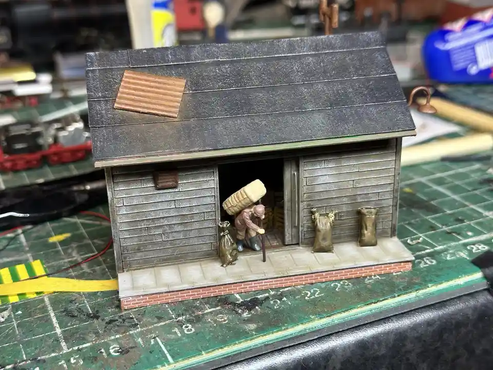 A miniature diorama of a rustic wooden shack with a dark roof and a small window, placed on a green cutting mat. A tiny…