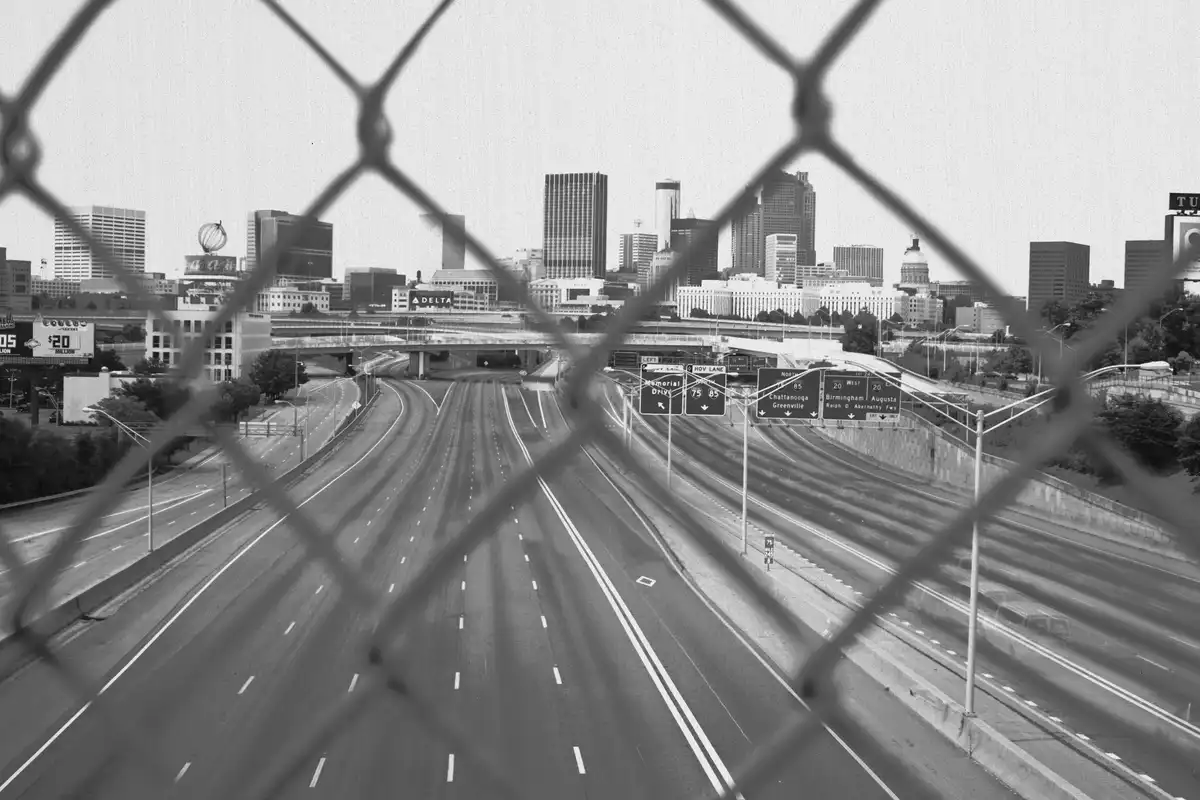 A black-and-white photo shows an empty highway with multiple lanes and road signs, viewed through a chain-link fence. The…