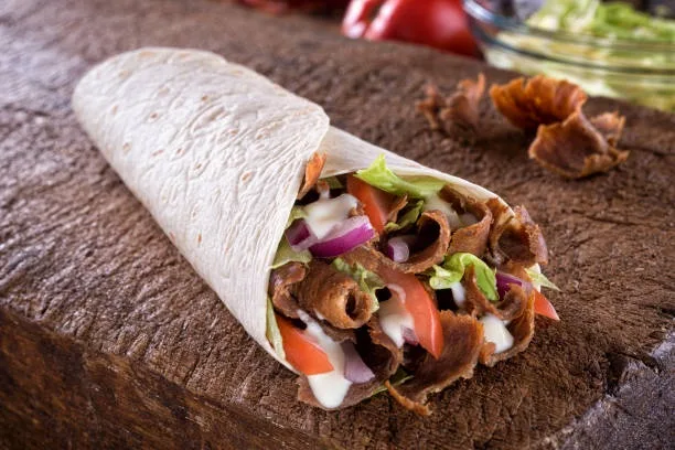 a close-up photo of a burrito wrapped in a flour tortilla, filled with meat, lettuce, tomato, onion, and sauce, resting on a…