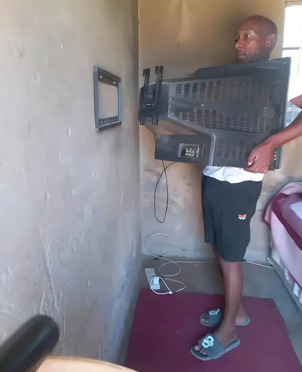 A man stands in a narrow room holding a large, bulky CRT television, its back facing the viewer, while another person's arm…