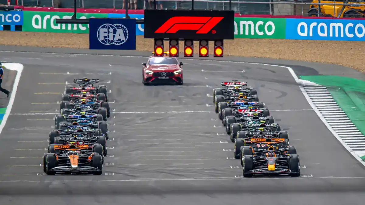 A wide shot of Formula 1 race cars lined up on a track, facing forward with a red safety car positioned ahead of them. Above…