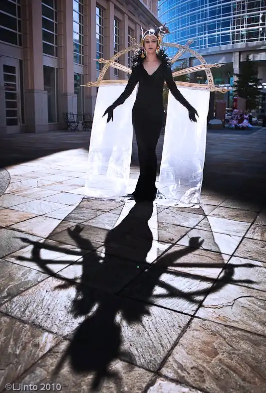A person in a black, elaborate costume with a large, ornate golden frame behind them stands in a sunlit plaza, casting a…