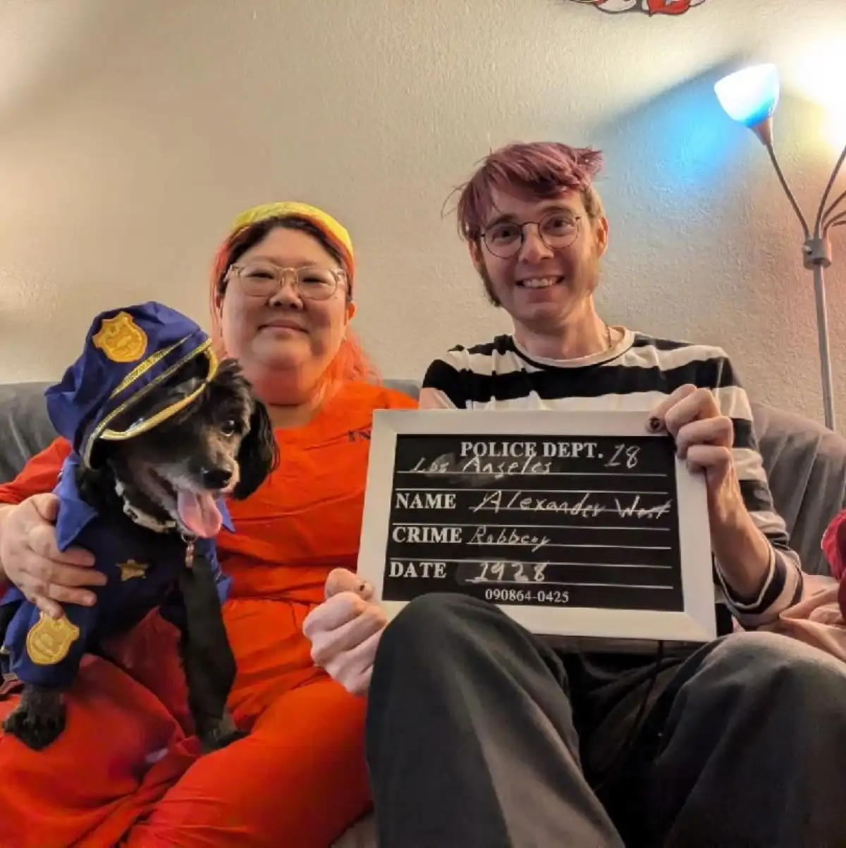 Two people and a small dog are posing on a couch. The woman wears an orange jumpsuit and holds a small black-and-white dog…
