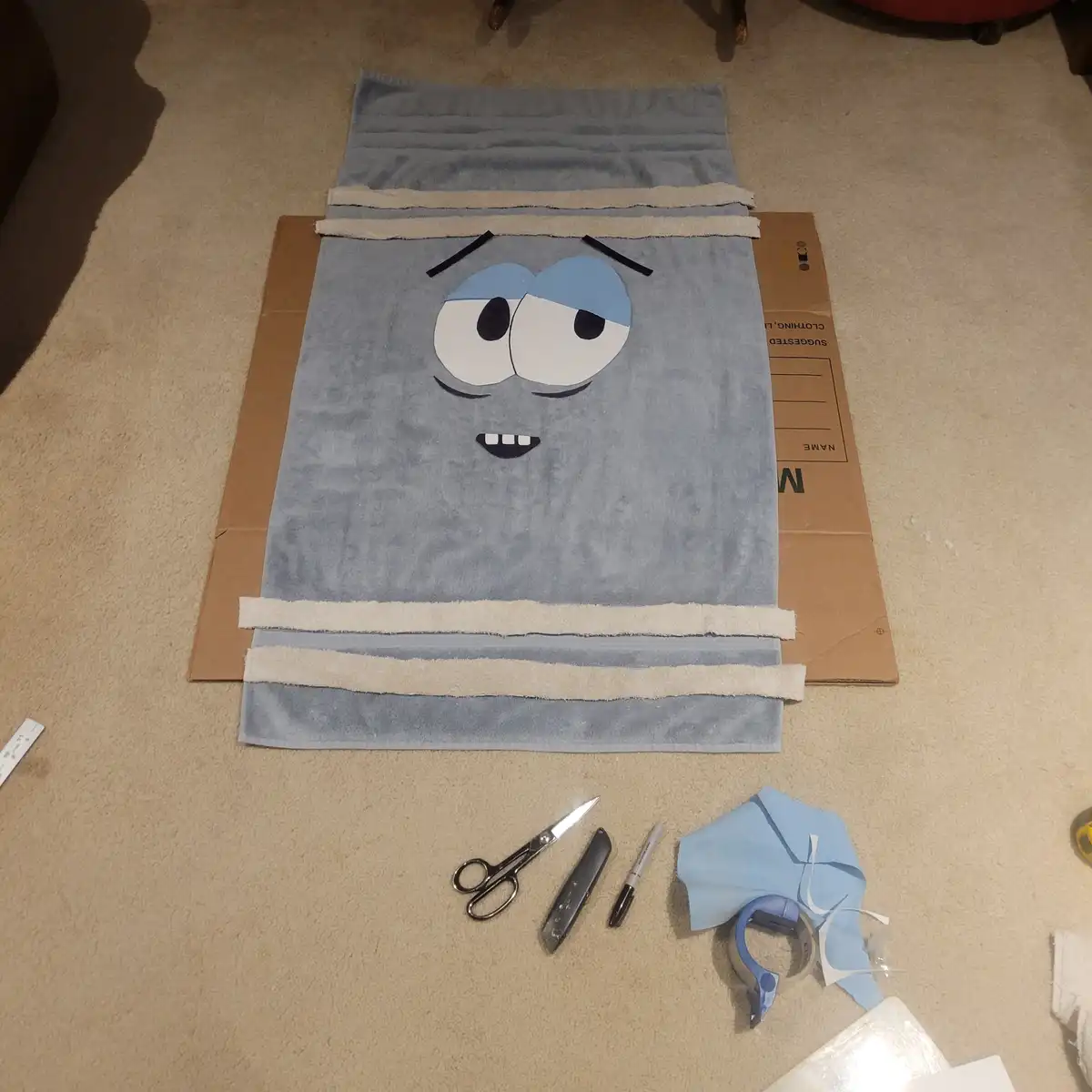 A light blue towel laid flat on a cardboard box on a beige carpet, with a cartoon face made of fabric pieces (large eyes…