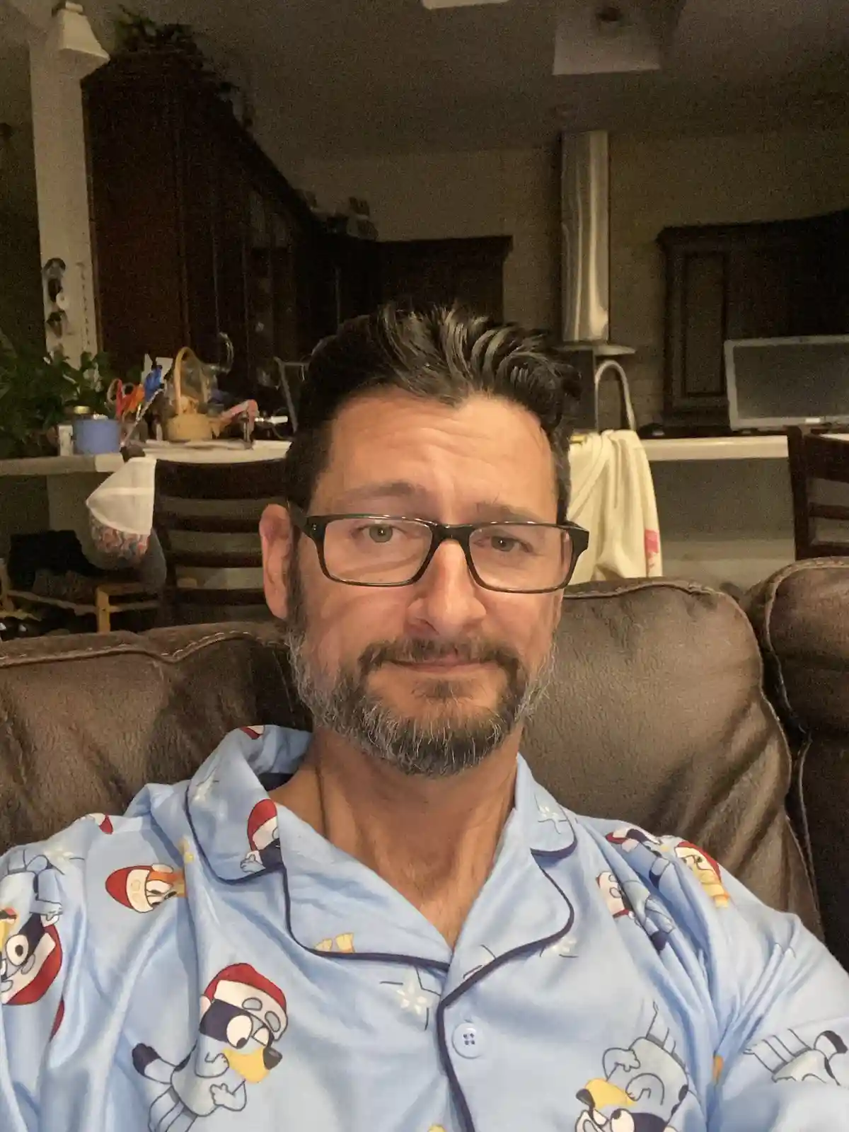A middle-aged man with glasses and a beard, wearing light blue pajamas with cartoon characters, sits on a brown couch in a…