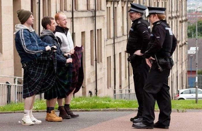 Three men in kilts stand facing two police officers on a sidewalk. The men appear to be explaining themselves, while the…