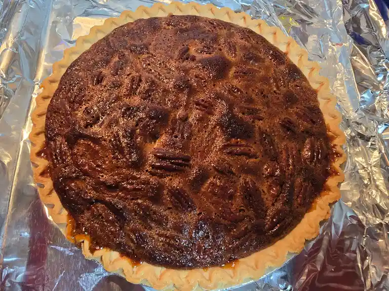 A close-up photo of a freshly baked pecan pie in a fluted crust, resting on crinkled aluminum foil. The pie's dark, glossy…
