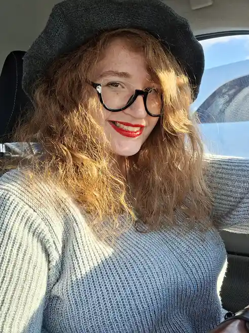 A woman with curly reddish-brown hair, wearing a black beret, large black-rimmed glasses, and bright red lipstick, smiles…