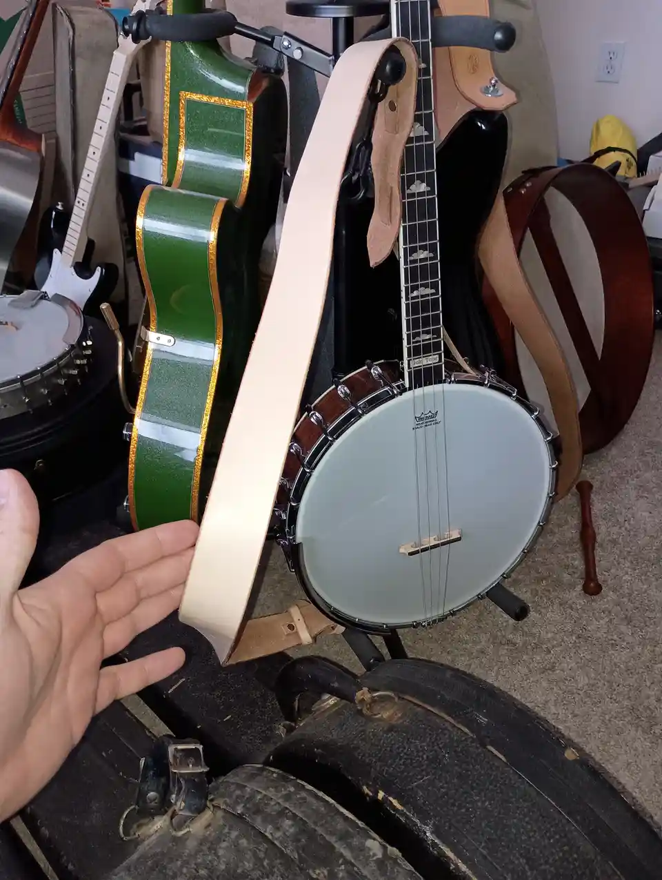 A person's hand holds up a light-colored leather strap next to a banjo mounted on a stand, with other string instruments…