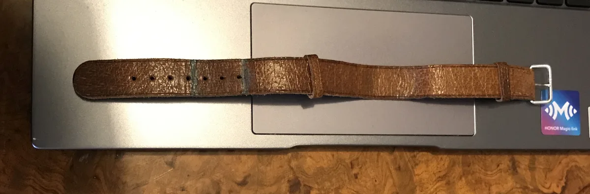 A brown leather watch strap with a silver buckle lies across the trackpad of a silver laptop. The strap has a noticeable…