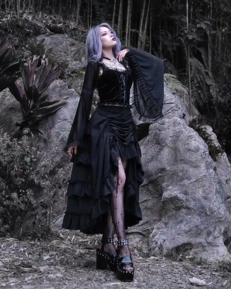 A person with lavender hair stands outdoors in a dark, gothic outfit featuring a black corset, layered ruffled skirt…