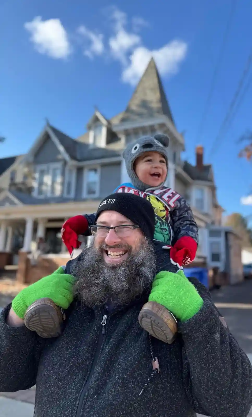A man with a long gray beard and glasses, wearing a black beanie and green gloves, carries a smiling toddler on his…