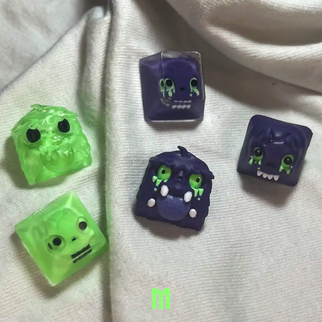 Five custom keyboard keycaps with cartoonish monster faces are arranged on a white fabric surface. Four are purple with…
