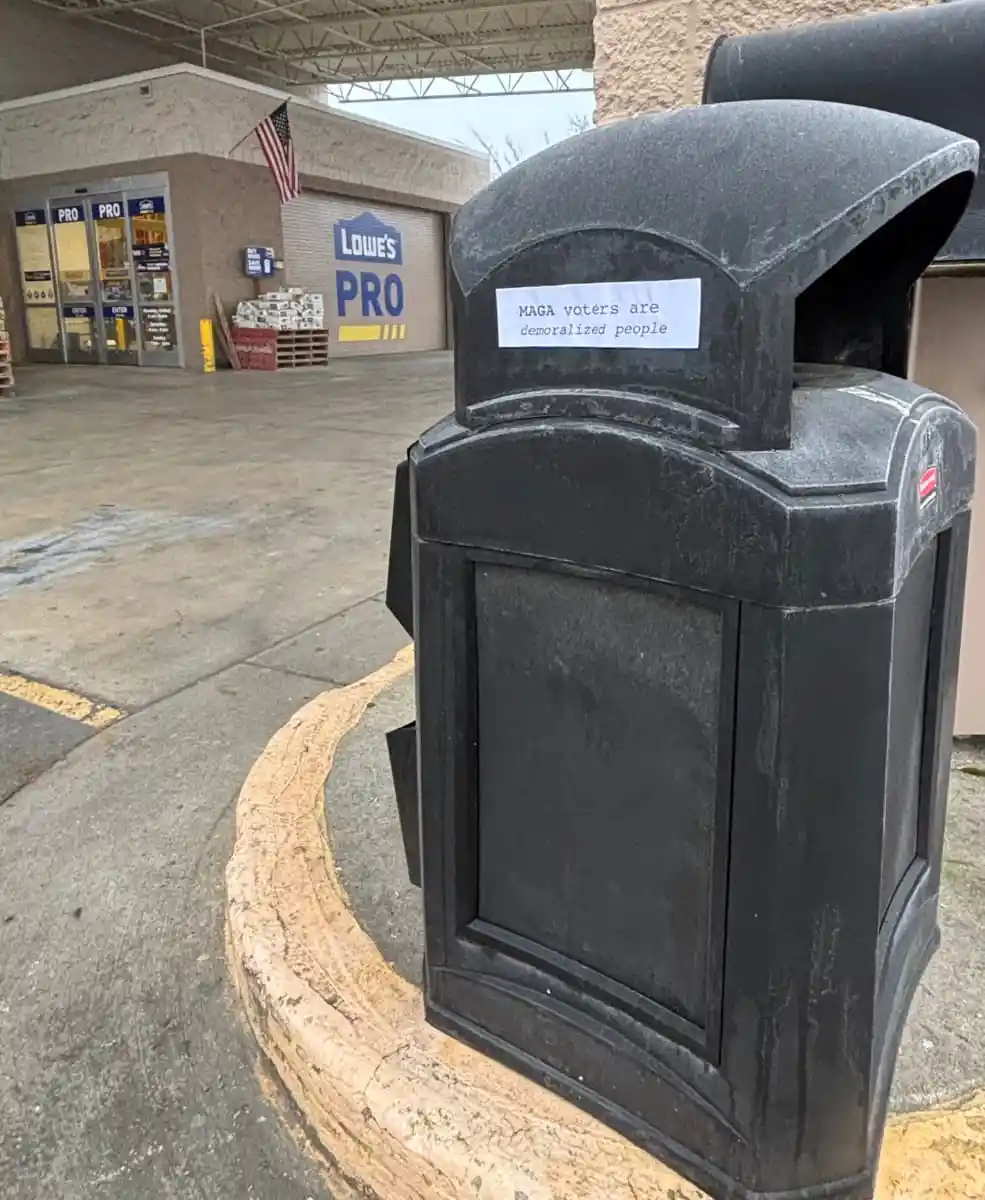 A black public trash can with a white paper note taped to its top reads 'MAGA voters are demoralized people'. The can is…