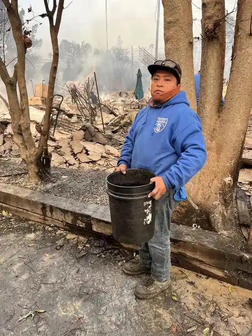 A man in a blue hoodie and cap stands in front of a burned-out property holding an empty black bucket, looking solemnly at…