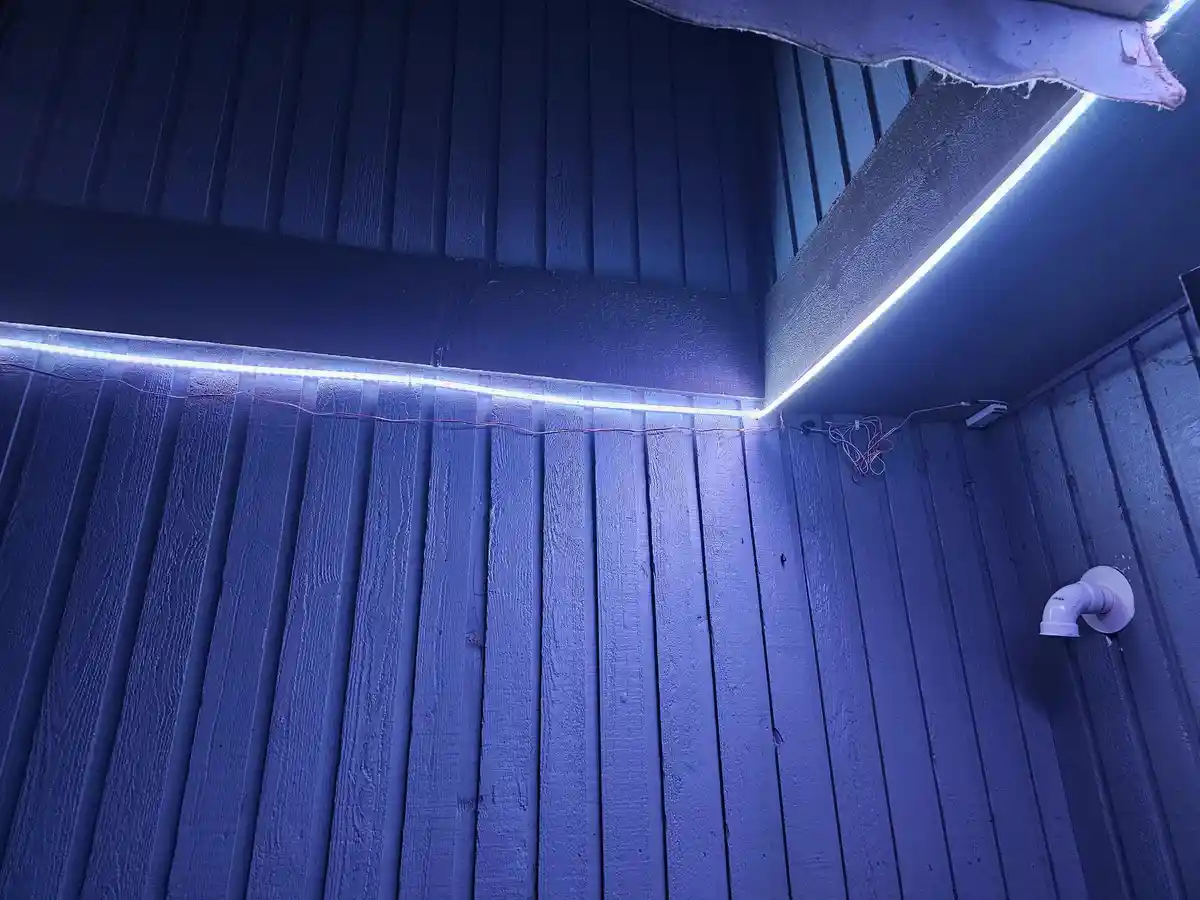 A photo shows bright white LED strip lights installed along the corner where a dark blue wooden wall meets a ceiling…