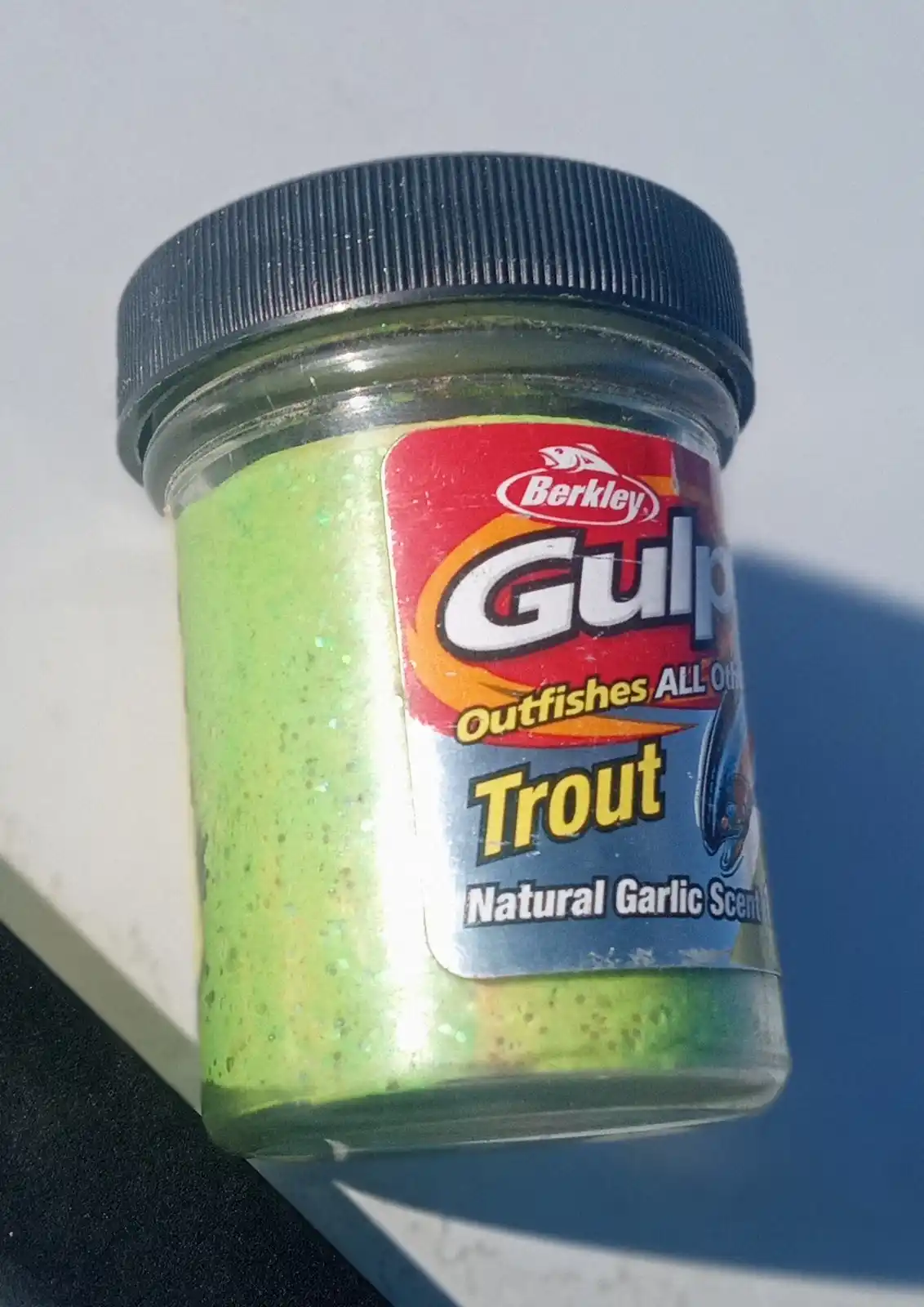 A close-up photo of a small glass jar with a black lid, filled with bright green, glittery fishing bait. The label reads…