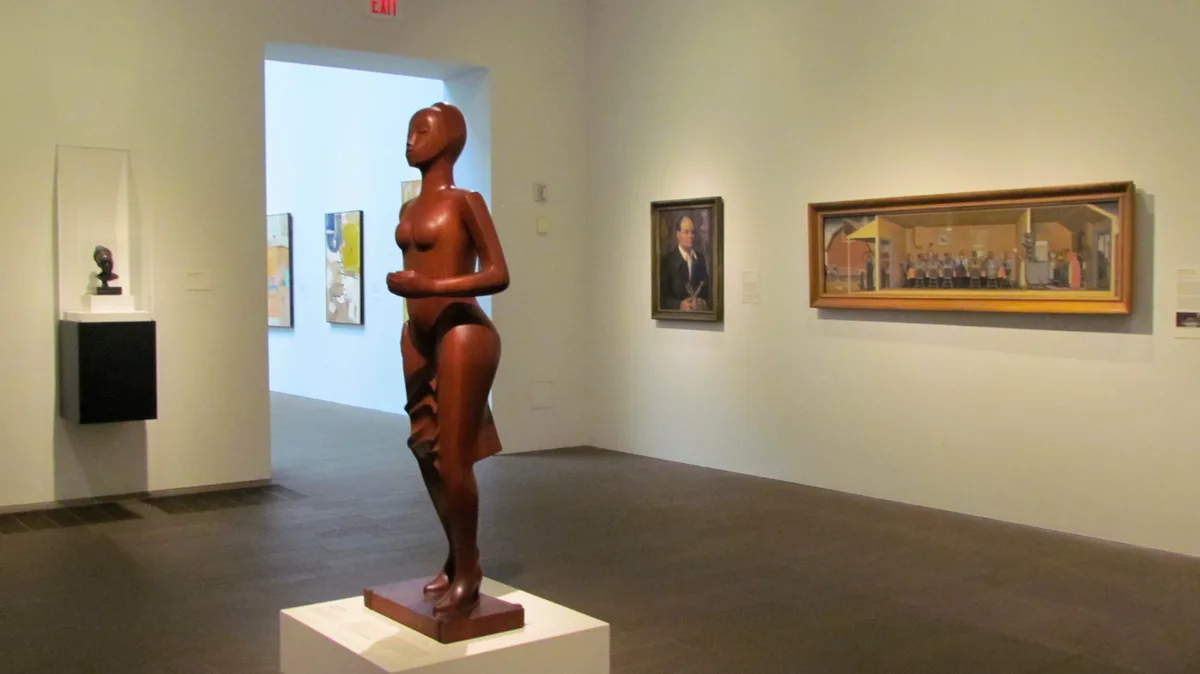 A polished wooden sculpture of a nude female figure stands on a pedestal in a modern art gallery, surrounded by other…