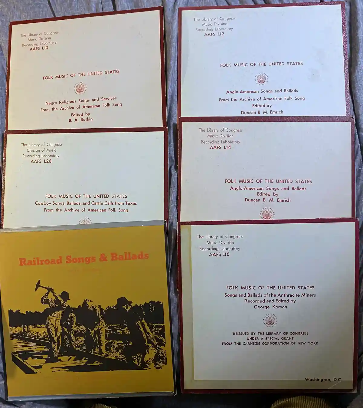 Five vintage Library of Congress folk music record albums are arranged on a fabric surface. Four have cream-colored covers…