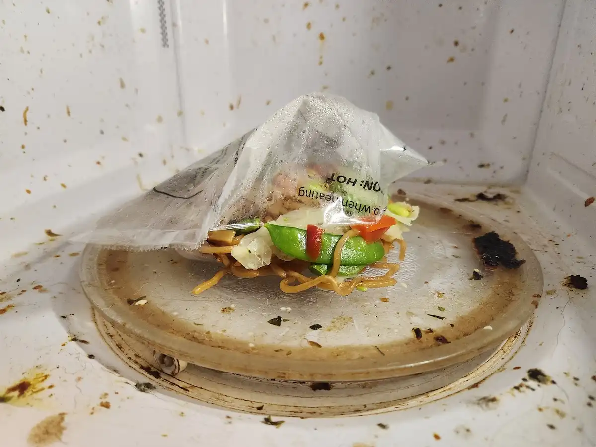 A close-up photo shows a heavily splattered and dirty microwave interior with a partially opened, steamy plastic food bag…
