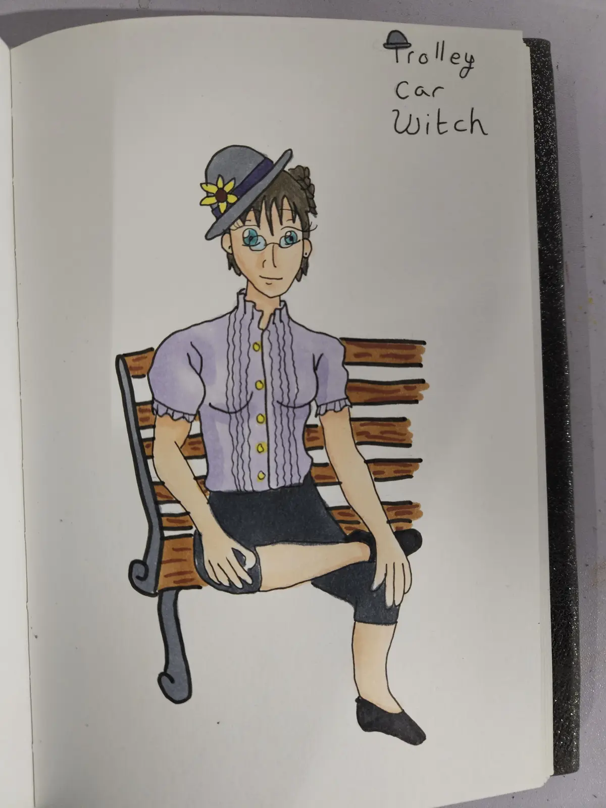 A hand-drawn illustration in a notebook shows a woman with short brown hair, glasses, and a grey hat with a yellow flower…