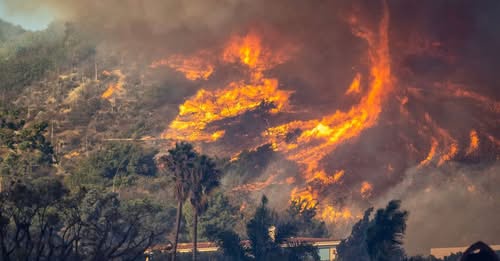 A dramatic photograph shows a large wildfire engulfing a hillside, with bright orange flames and thick smoke billowing…