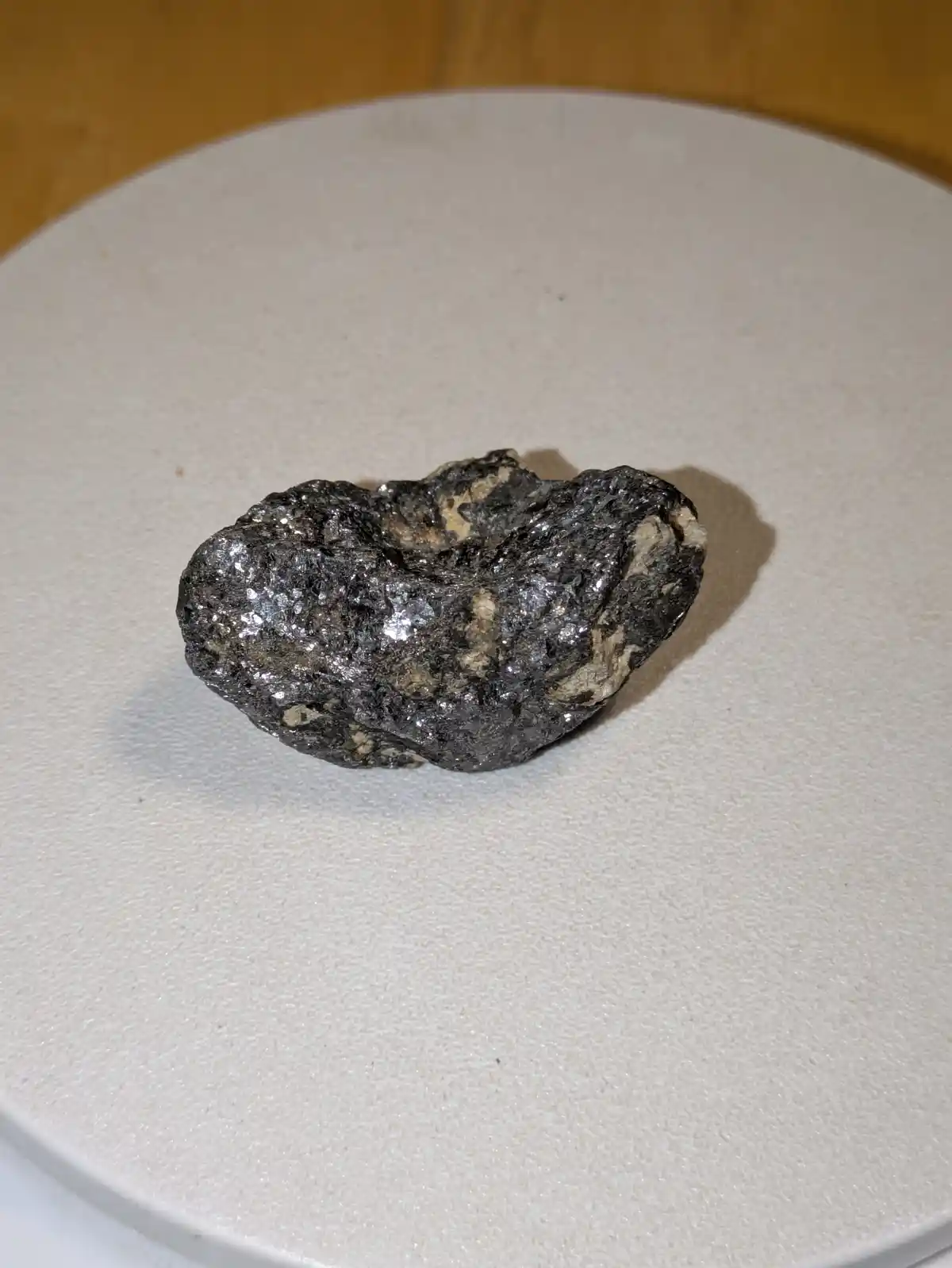 A close-up photo shows a rough, irregularly shaped rock with a metallic sheen and speckled gray and black texture, resting…