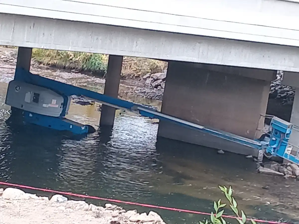 A blue truck is flipped upside down and partially submerged under a concrete bridge over a river, with its rear end resting…