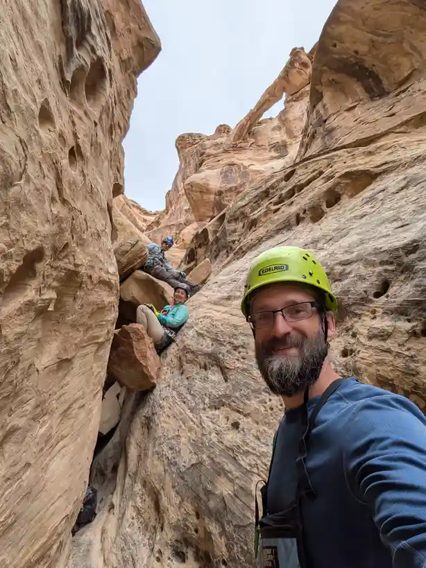 A selfie of a man with a green Edelrid helmet and glasses smiling at the camera inside a narrow slot canyon with textured…