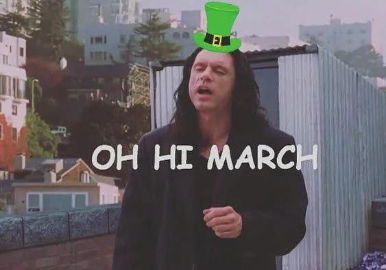 A man with long dark hair and a black coat stands outdoors with a green leprechaun hat digitally placed on his head. The…