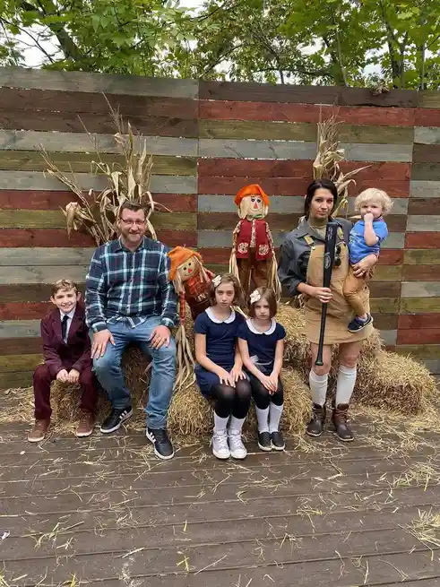 A family of five poses for a photo in front of a rustic wooden wall decorated with corn stalks and scarecrows. The father…