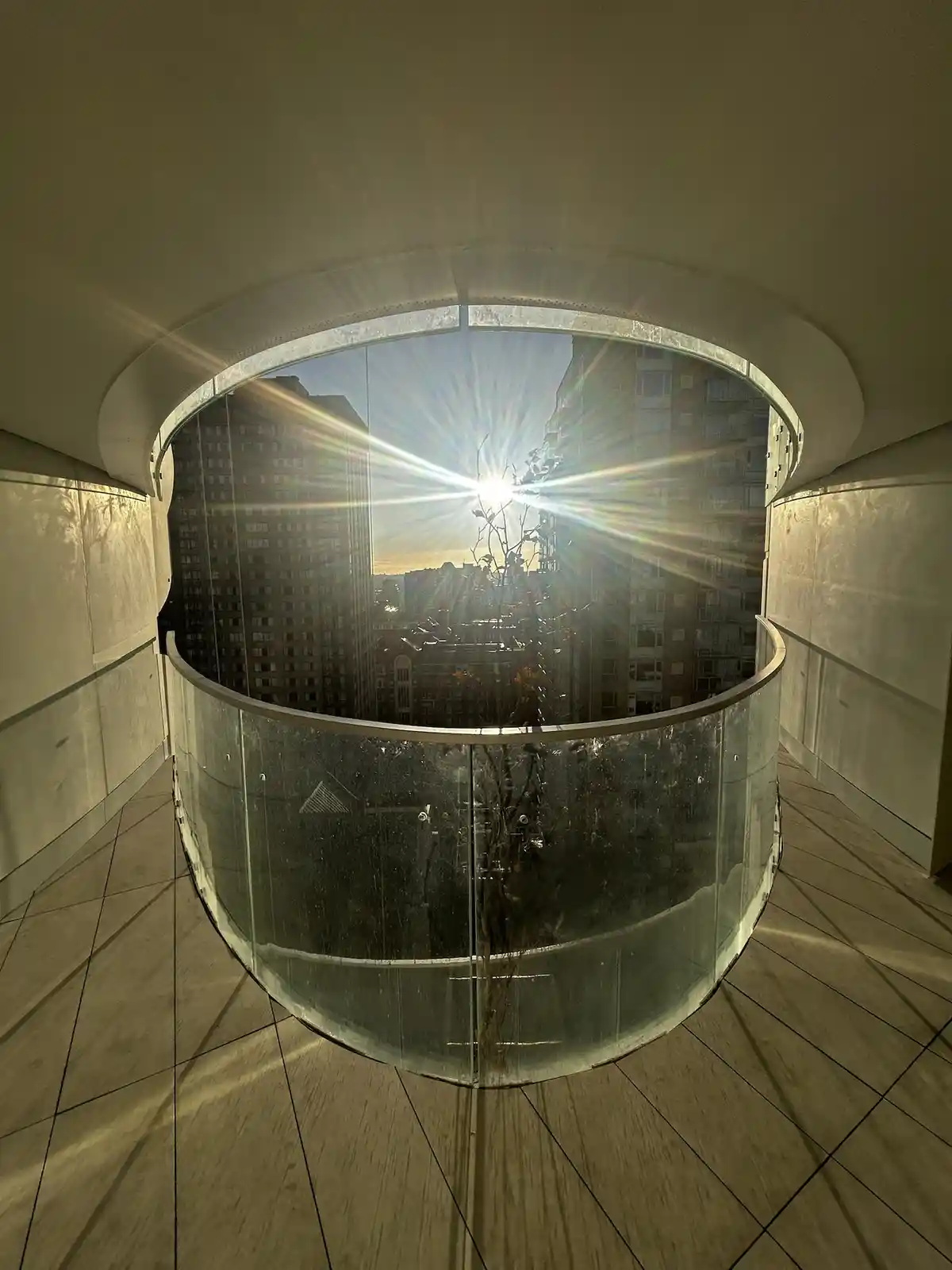 A photo of a curved glass balcony with a cityscape view, where the sun is shining brightly through the glass, creating a…