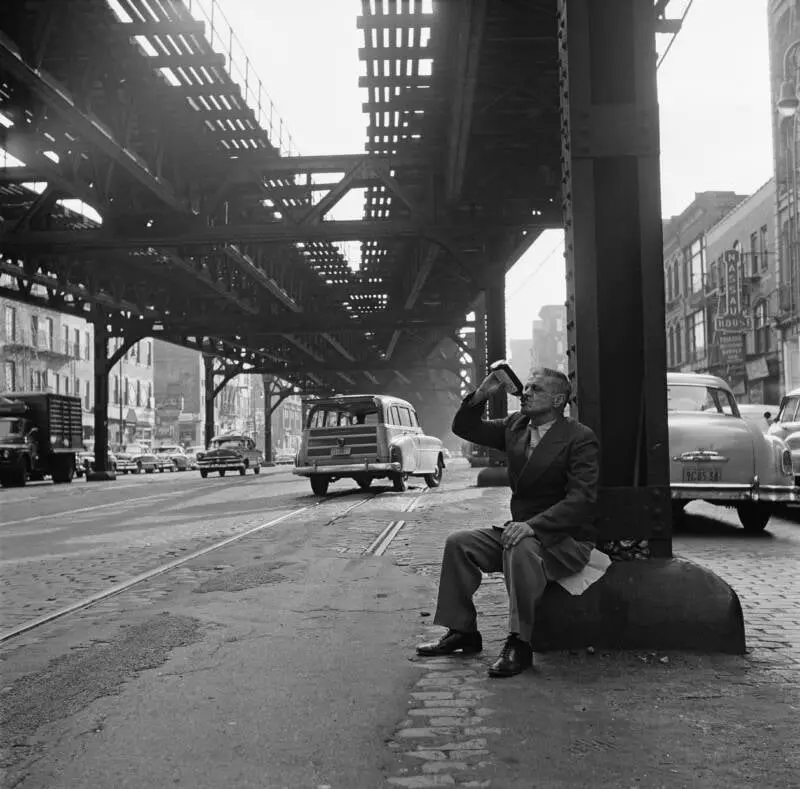 a black-and-white photograph shows a man in a suit sitting on a large metal support pillar under an elevated train track…