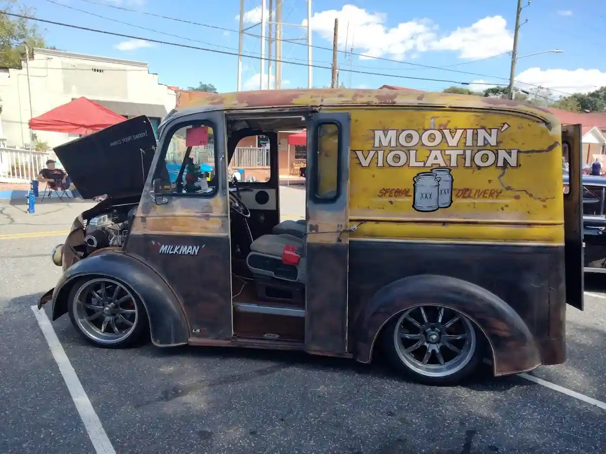 A rusted, customized vintage milk truck with its hood open and doors ajar, parked in a lot. The side reads 'MOOVIN'…