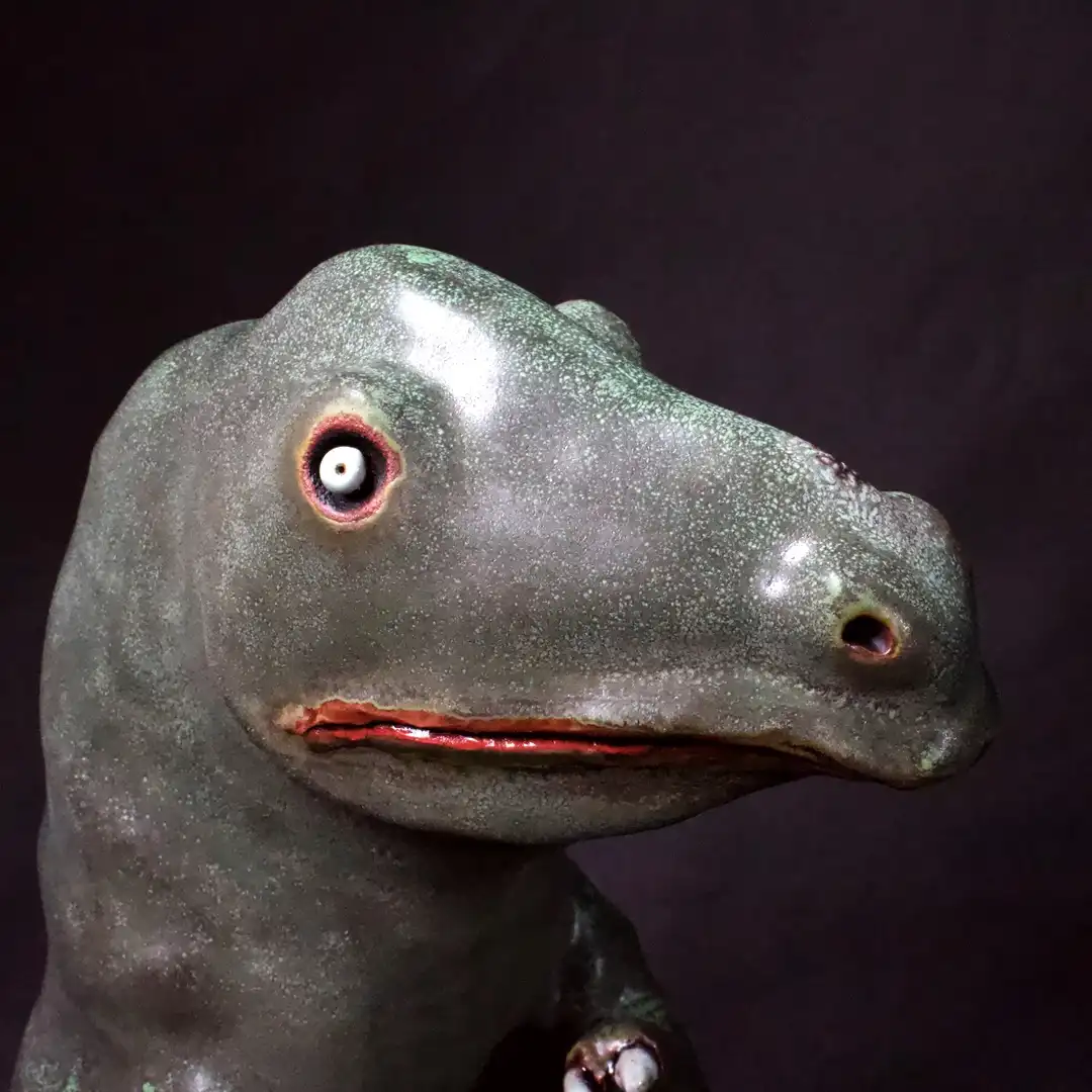 A close-up of a gray, textured, toy-like dinosaur head with one large, white, cartoonish eye and red lips, set against a…