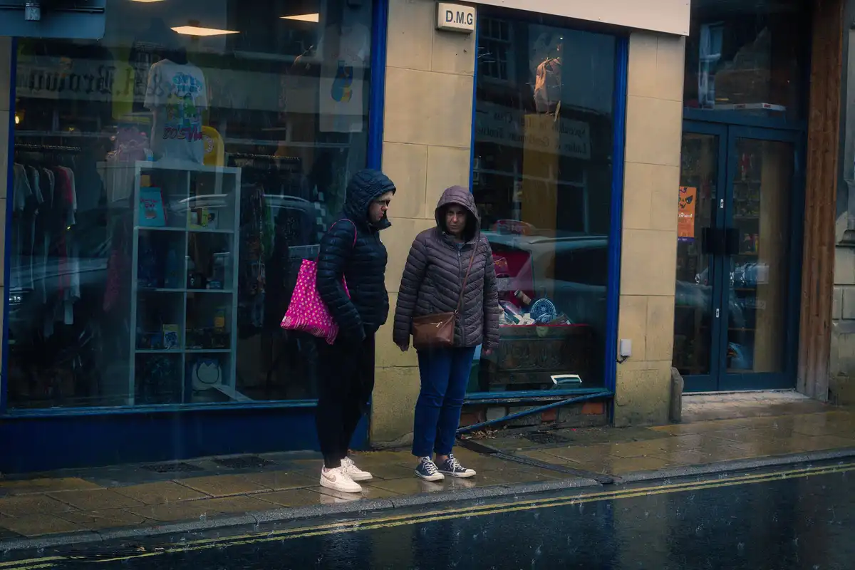 Two women stand on a wet sidewalk outside a shop during rain, both wearing hooded jackets and looking at each other with…